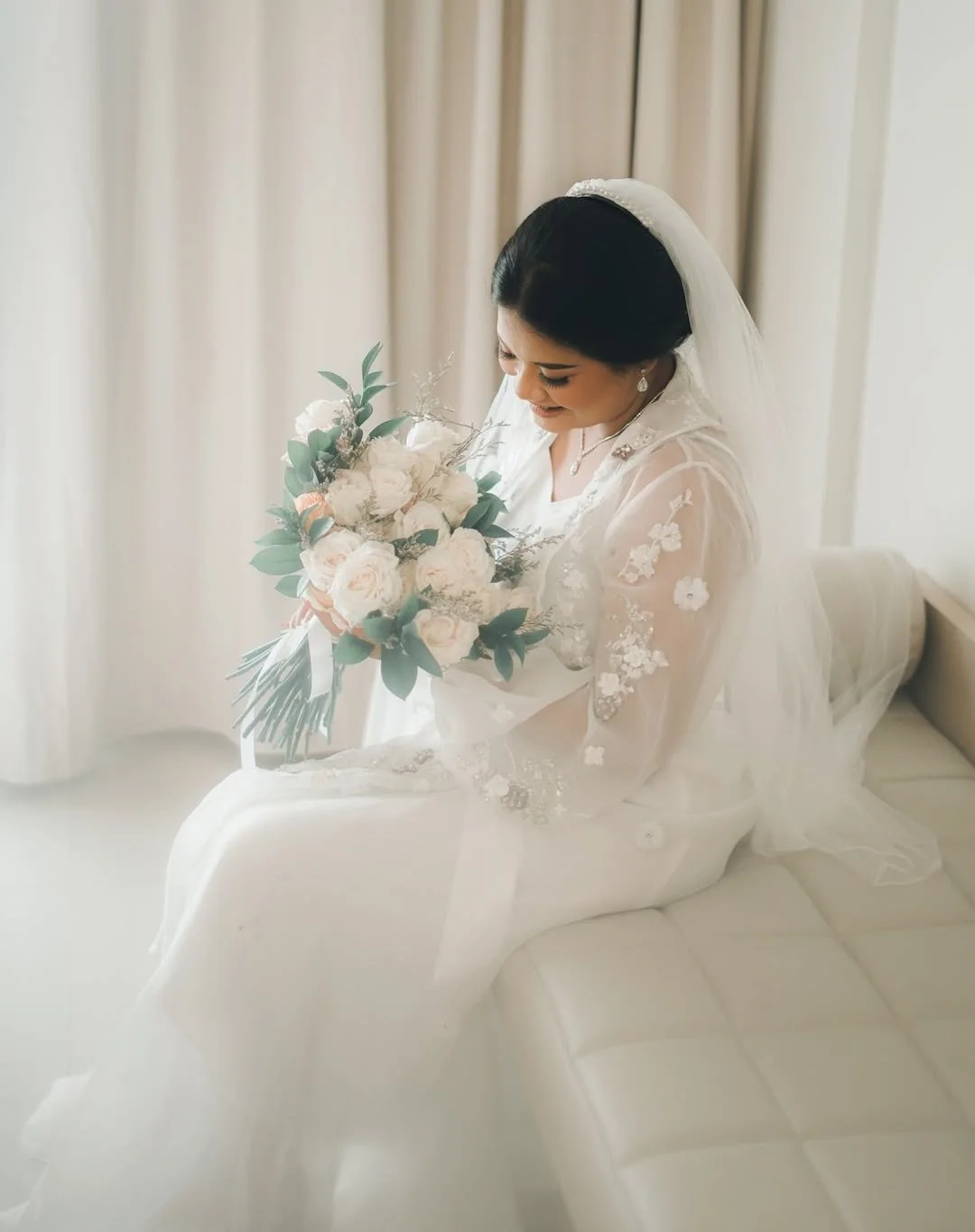 Bride in a white dress holding a bouquet beside a window