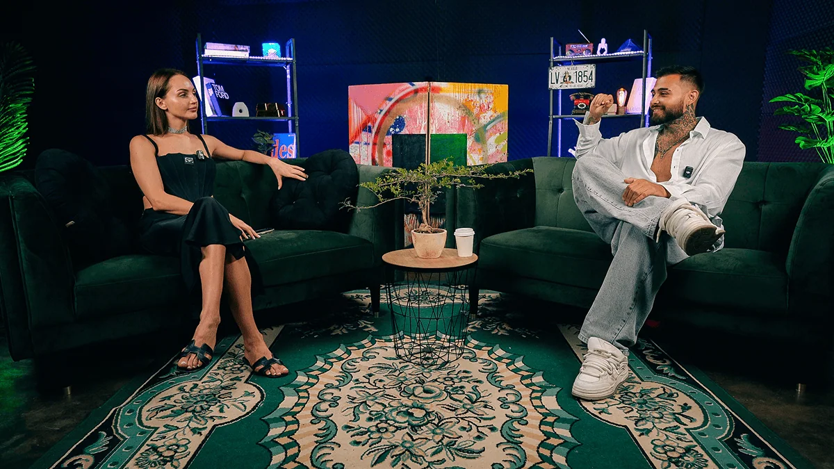 Relaxed studio interview setting with two people on plush green sofas.