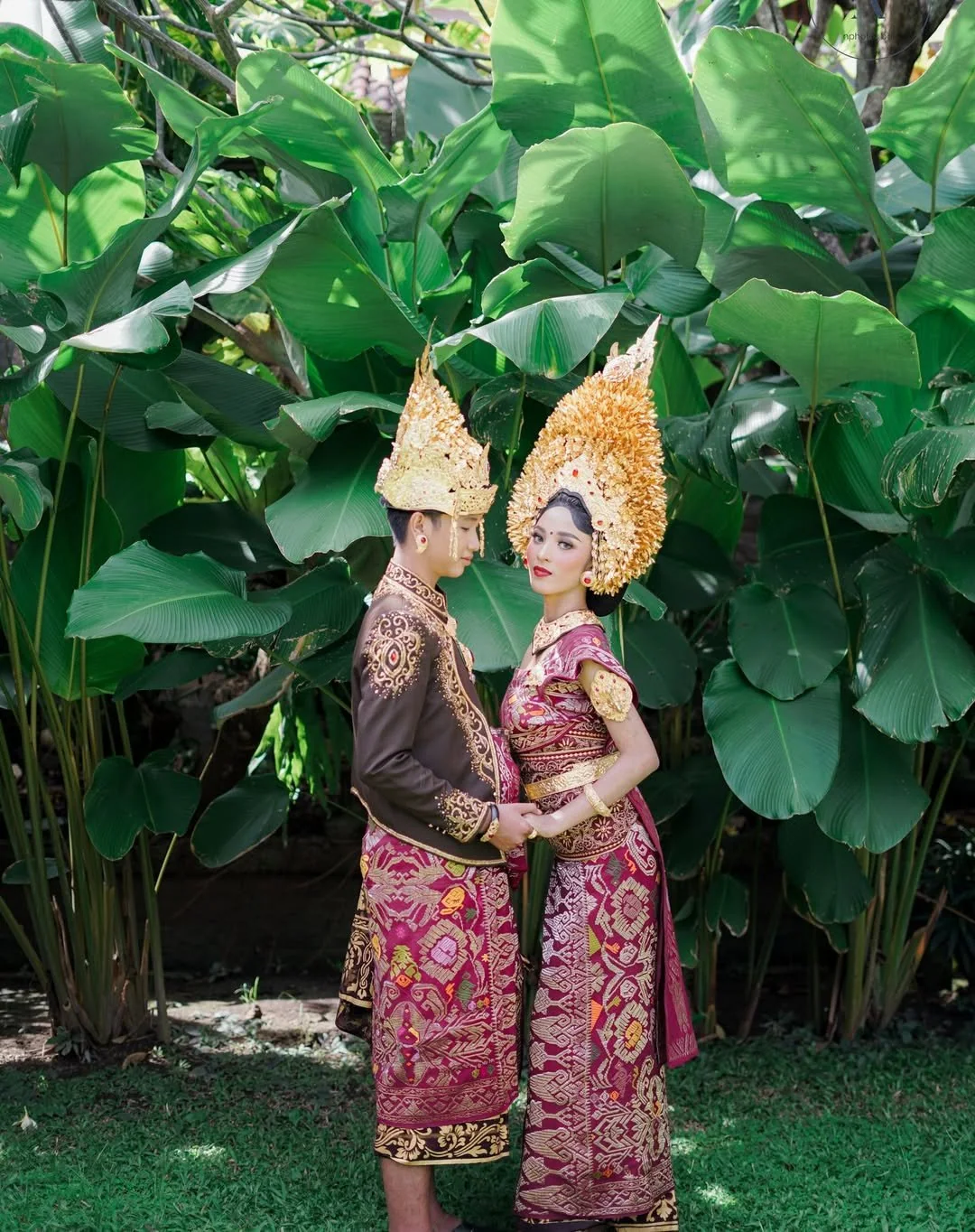 Couple in traditional Balinese wedding clothes standing among green leaves