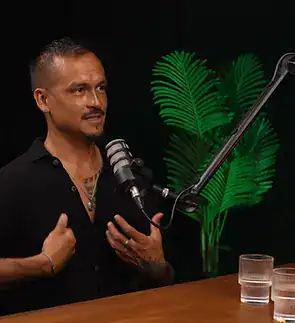 A man speaking into a microphone during a podcast or interview session with plants in the background.